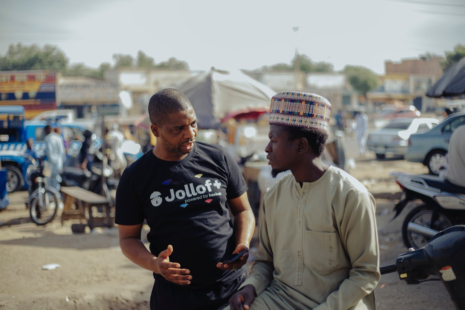 Two men talking at a busy roadside market, one holding a phone