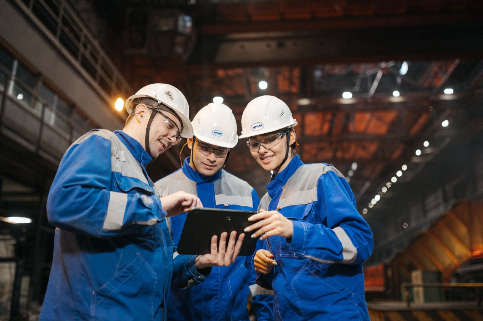 Three engineers in hard hats reviewing production data on a tablet beside a furnace